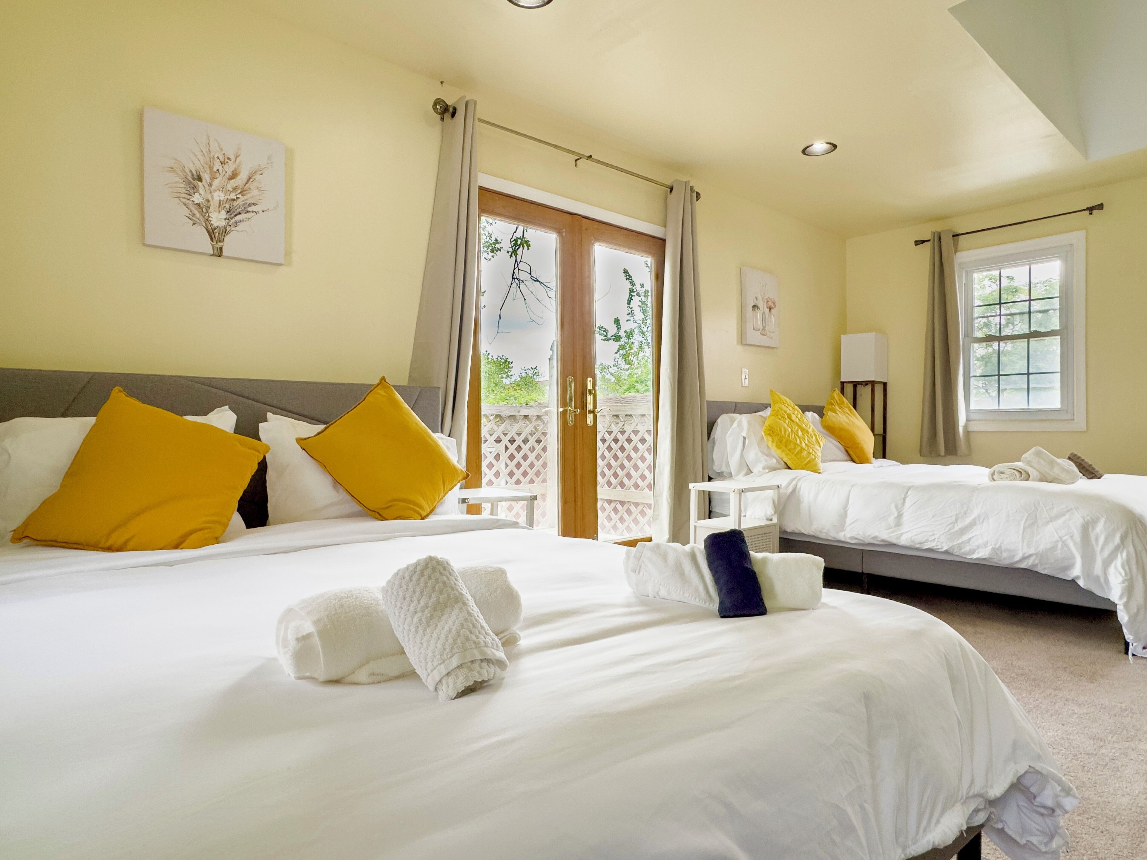 A hotel room with two beds, each with white bedding and yellow pillows. There are towels on the beds, and a window with curtains.