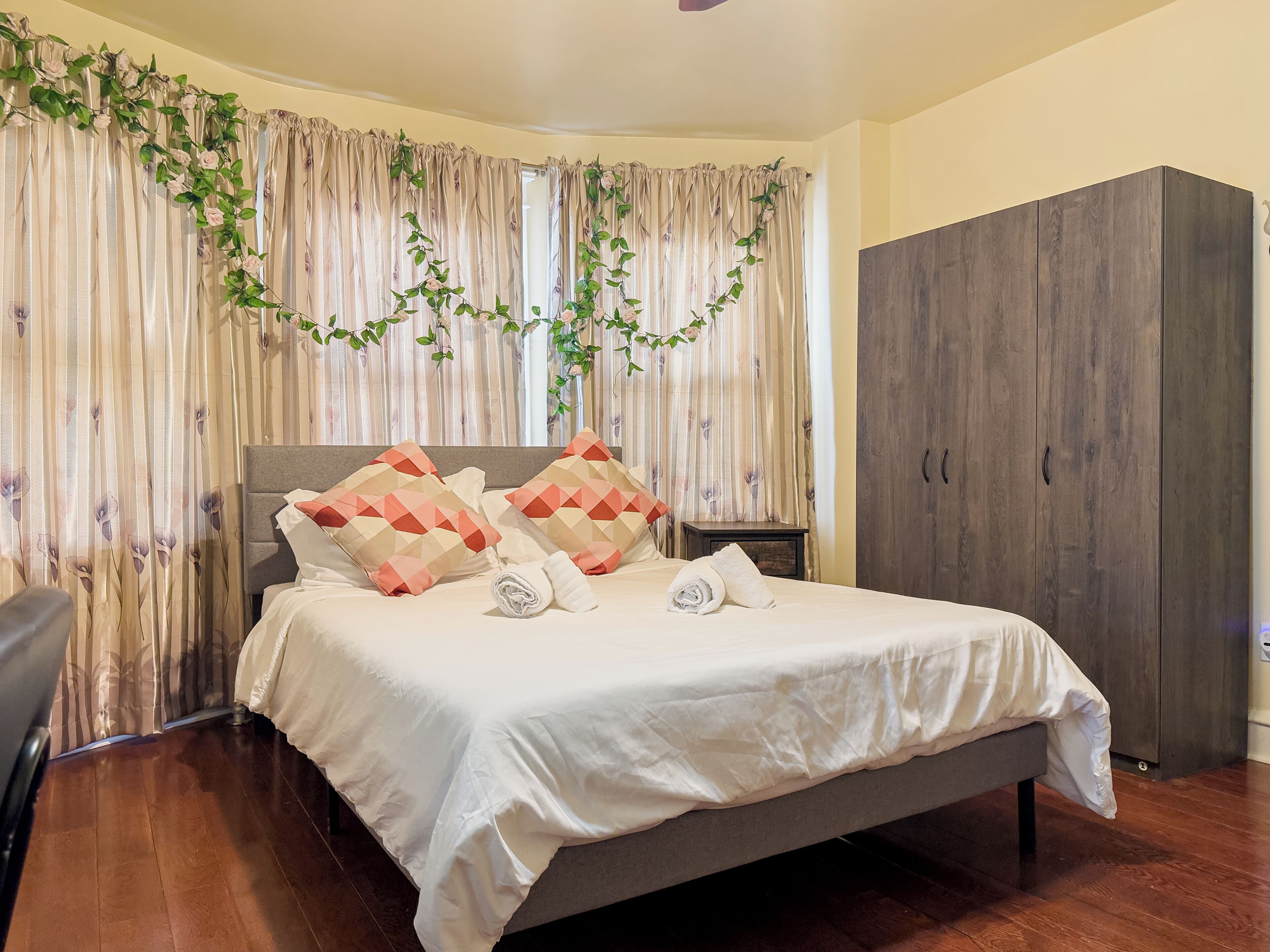 Een opgemaakt bed met een grijze hoofdbord, twee kussens en twee opgerolde handdoeken. Aan de rechterkant staat een houten garderobe en een raam met gordijnen en groene klimplanten.
