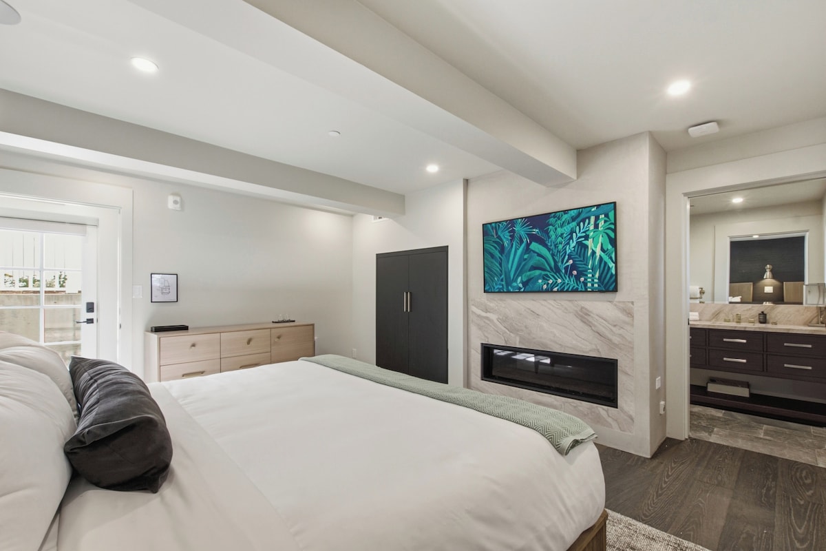Un dormitorio moderno con una cama, un aparador, un televisor montado en la pared y un baño con espejo y lavamanos.