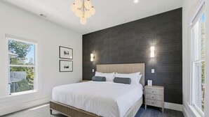 A modern bedroom with a large bed, a nightstand, a wall-mounted light, and a window with a view of trees.