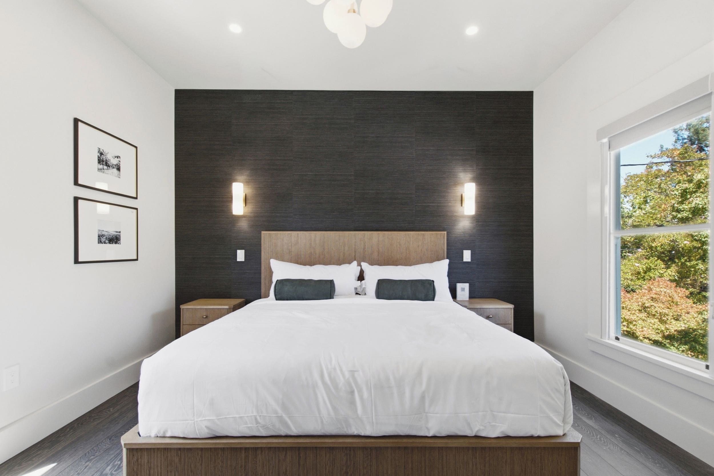 A modern bedroom with a large bed, dark wall paneling, two bedside tables, and a window with a view of trees.