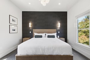 Un dormitorio moderno con una cama grande, paneles de pared oscuros, dos mesitas de noche y una ventana con vistas a árboles.