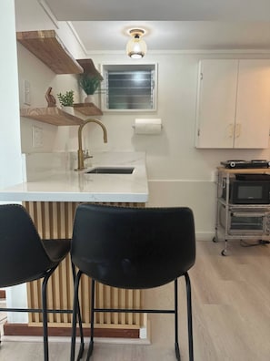 A compact kitchen with a white countertop, a gold faucet, and a wooden shelf. There are two black chairs and a white cabinet in the background. - Kaneohe Bay Ocean Views (Kaneohe)