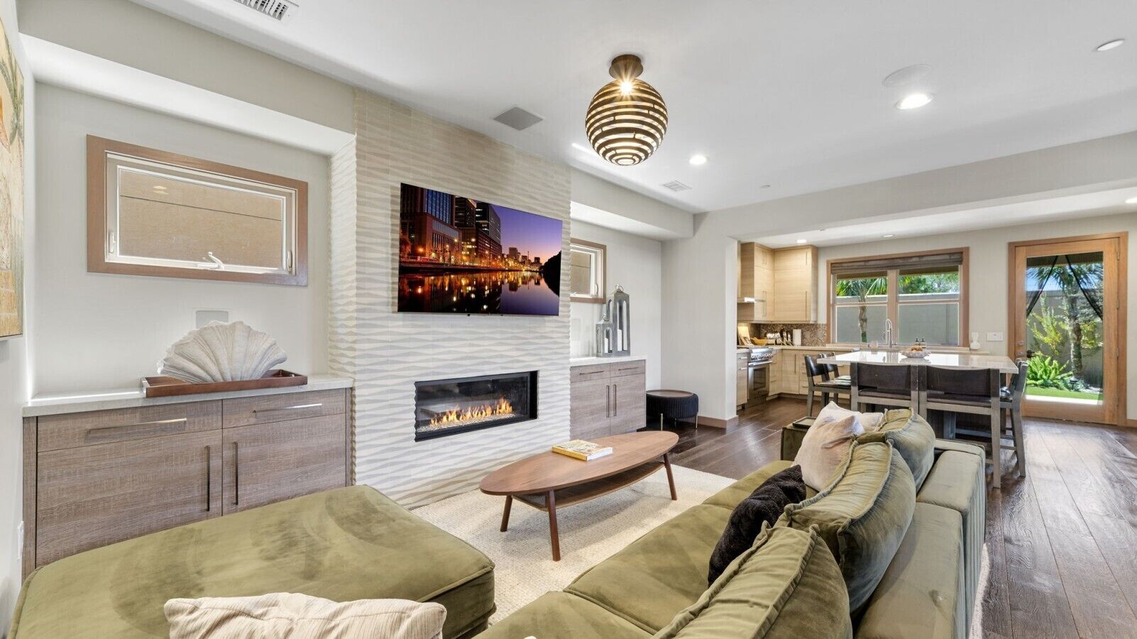 A modern living room with a fireplace, a flat-screen TV, and a dining area with a kitchen in the background.