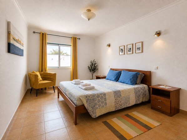 Ein Schlafzimmer mit einem Bett, einem Sessel, einem Nachttisch und einem Fenster mit Vorhängen.