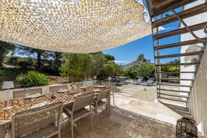 Un área de comedor al aire libre con una pérgola, una mesa preparada para comer, y vistas al jardín y la casa al fondo.