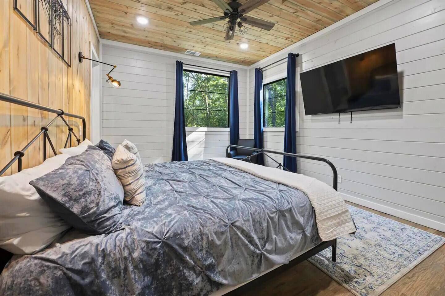 A bedroom with a bed, a ceiling fan, a TV mounted on the wall, and a window with curtains.