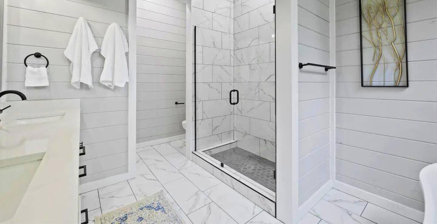 A bathroom with a shower, white tiles, a towel rack, and a rug.