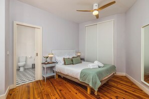 Een slaapkamer met een houten vloer, een bed met een wit hoofdeinde, een nachtkastje met een lamp en een plafondventilator.