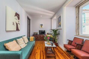 Een woonkamer met een blauwgroene bank, een rode fauteuil, een houten salontafel en een televisie achterin.