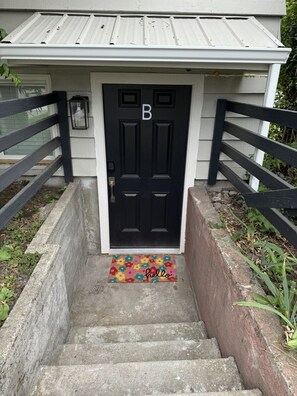 Uma porta preta com a letra 'B' acima, uma escadaria de concreto que leva à porta e um tapete de entrada colorido com a palavra 'hello'.