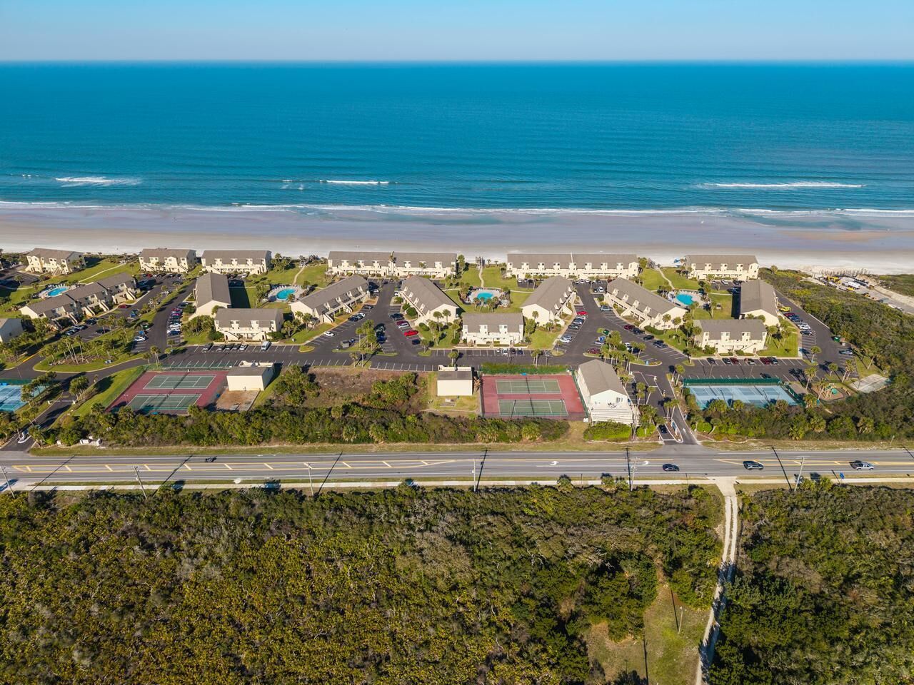An aerial view of a coastal resort with multiple buildings, a parking lot, and a road leading to the beach.