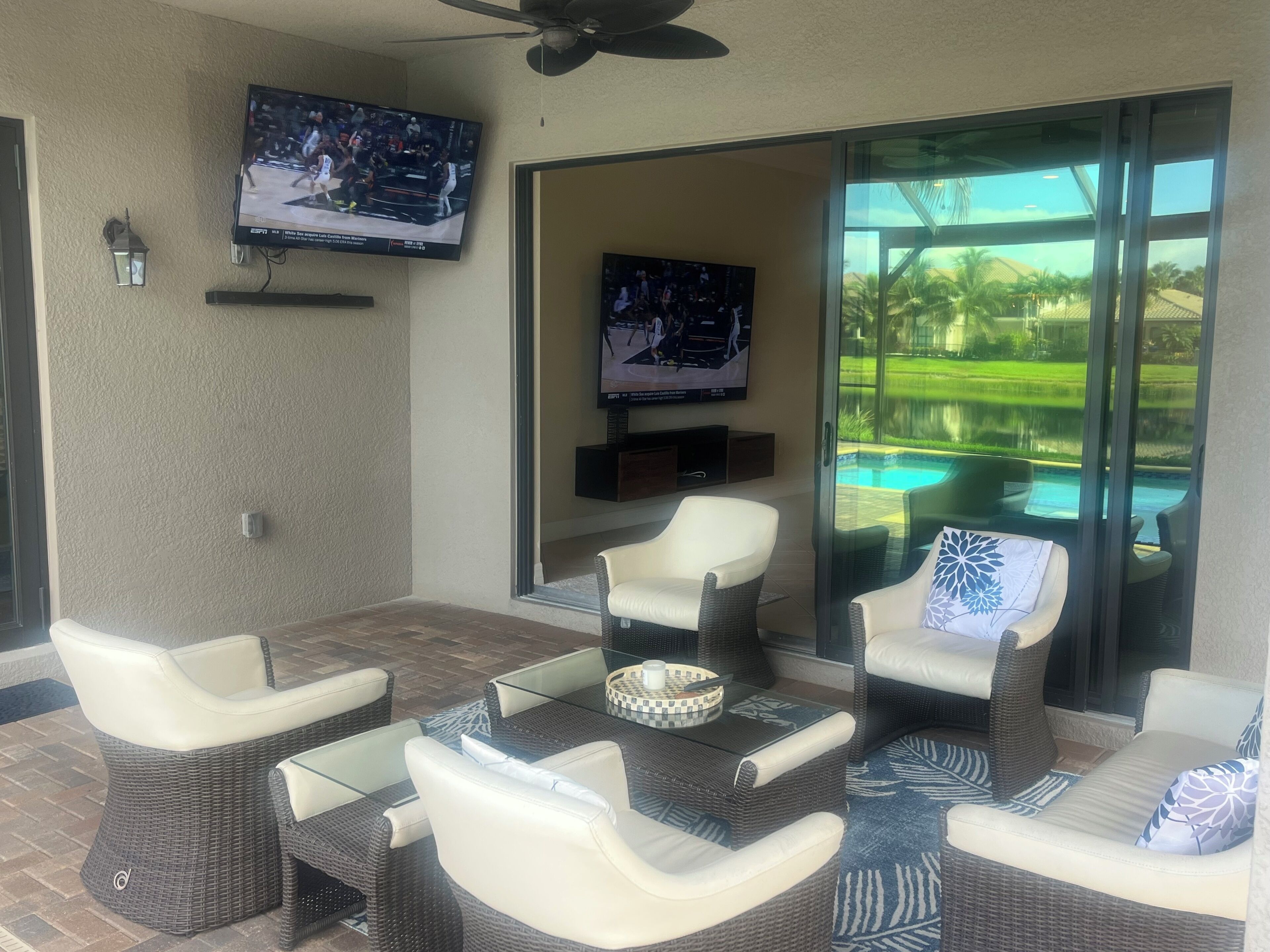 A patio area with wicker furniture, a flat-screen TV mounted on the wall, and a view of a swimming pool through large glass doors.