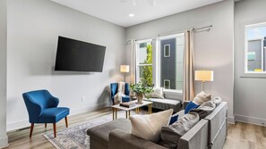 Ein modernes Wohnzimmer mit einem Flachbildfernseher, einem blauen Sessel, einem grauen Sofa und einem kleinen Tisch mit einer Pflanze.
