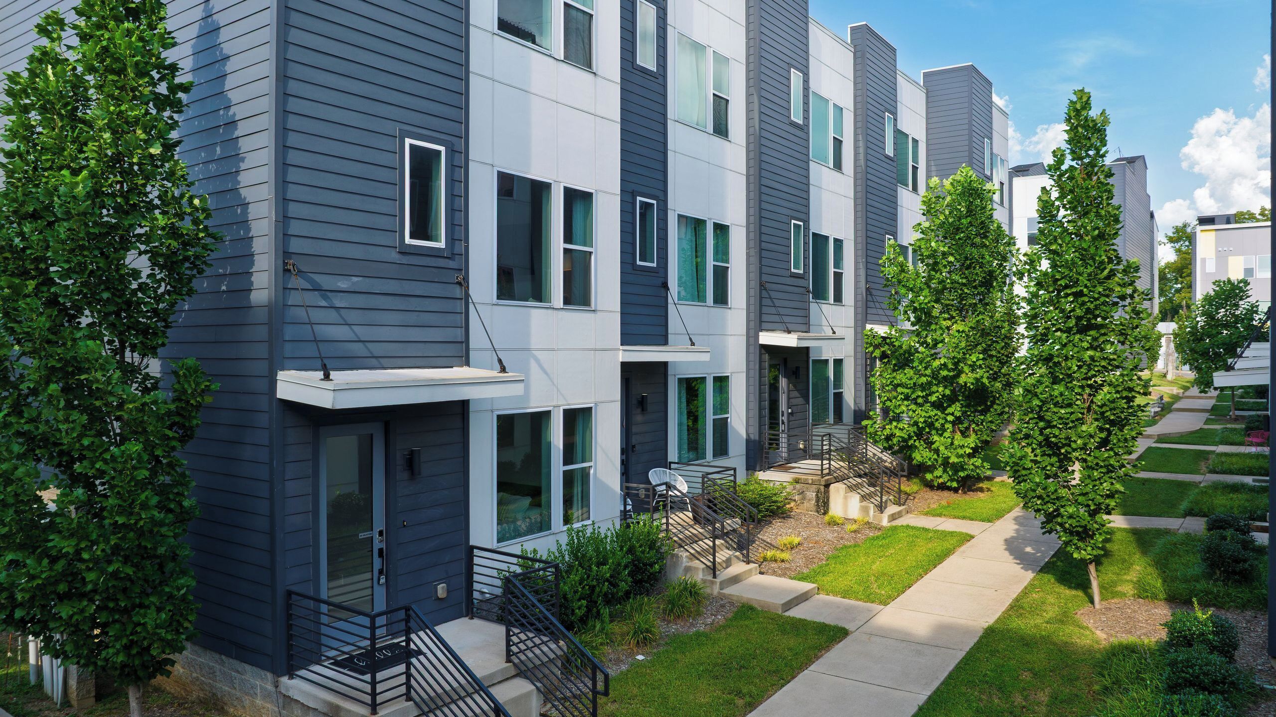 Un immeuble résidentiel moderne à plusieurs étages, doté d’un toit plat, de grandes fenêtres et d’un petit jardin devant, avec un chemin.