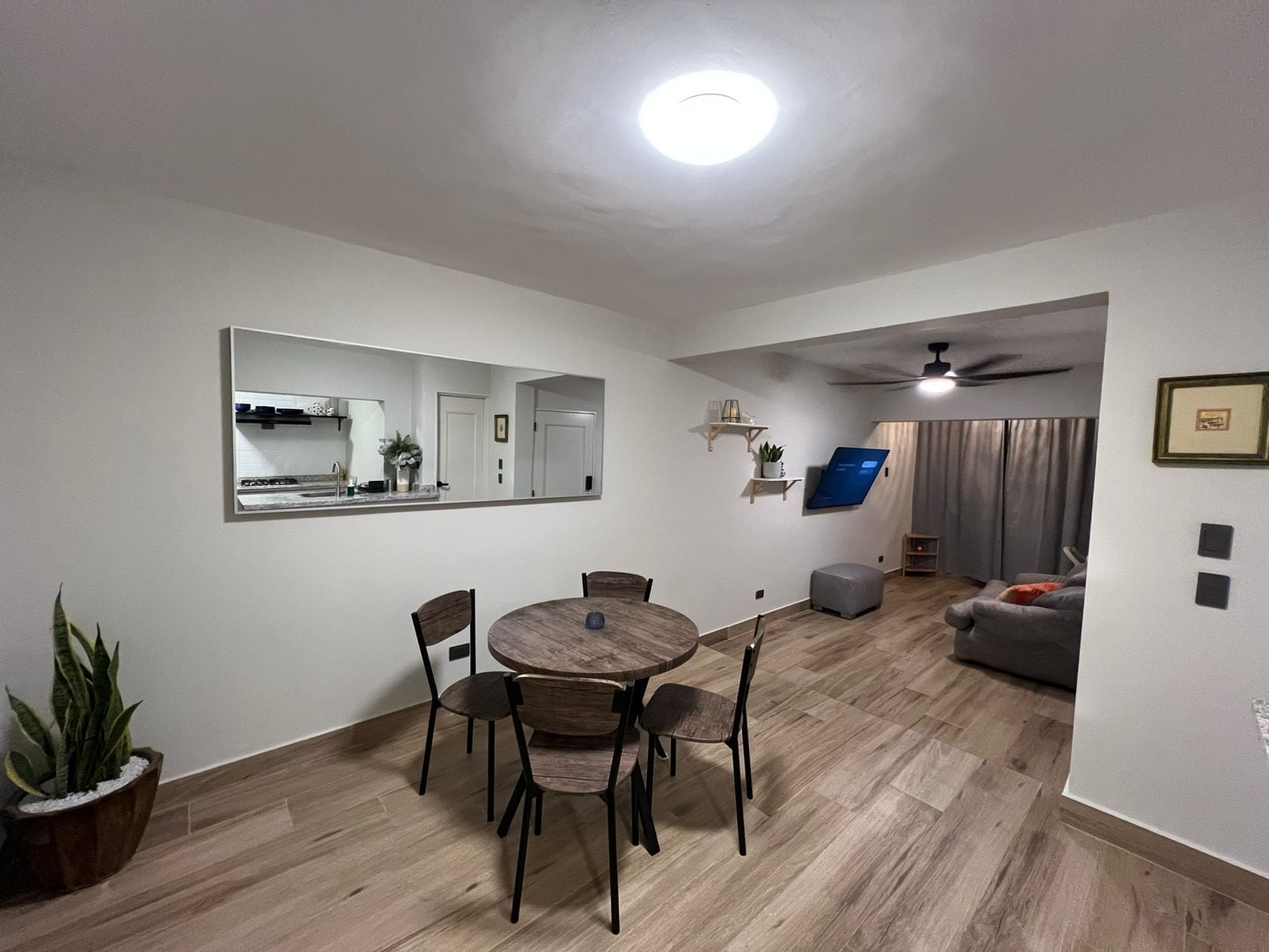 Ein Essbereich mit rundem Tisch und Stühlen, eine Küche mit Spiegel und eine Wohnfläche mit Sofa und Vorhängen.