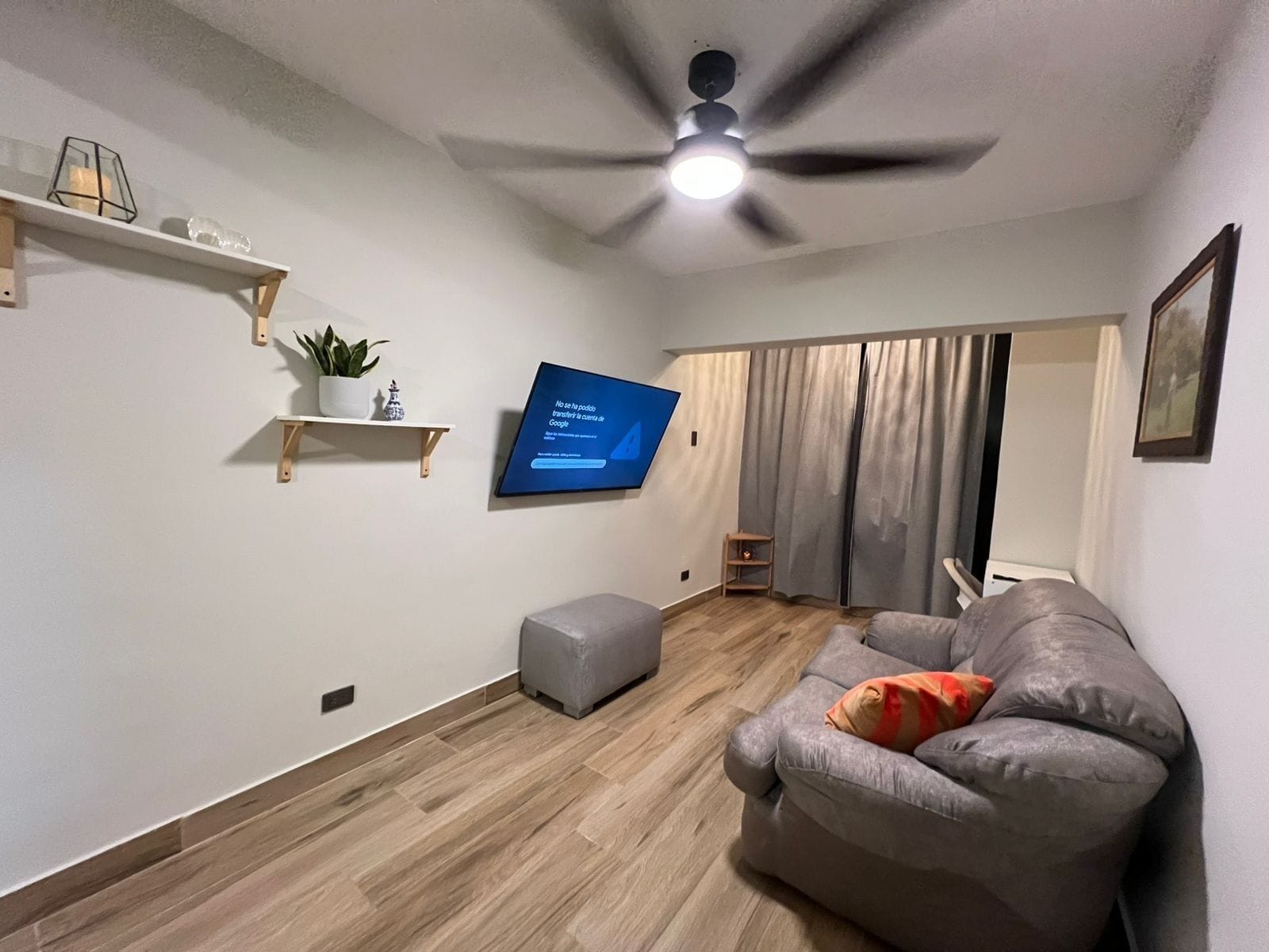 Ein Wohnzimmer mit Deckenventilator, einem an der Wand befestigten Flachbildfernseher, einem grauen Sofa mit Kissen, Holzboden und einem kleinen Holzregal mit Pflanze.