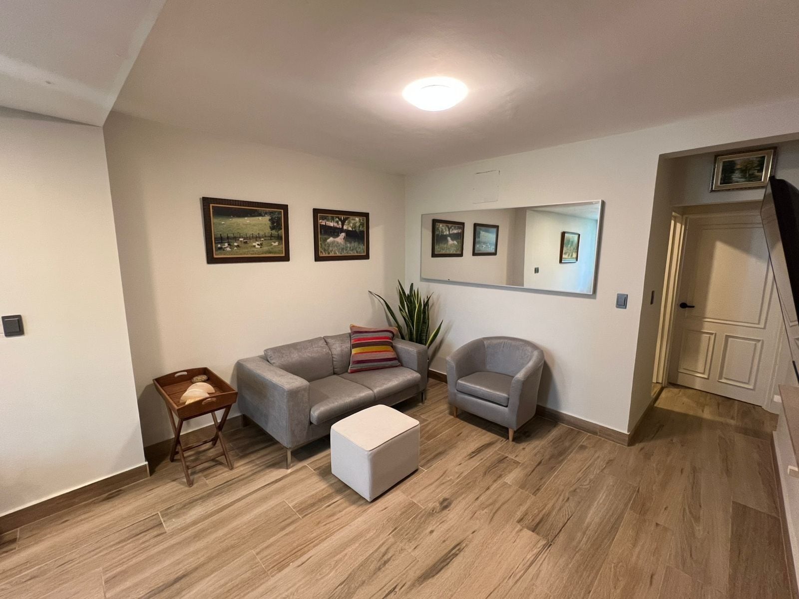 A living room with a grey sofa, a white ottoman, a small wooden side table, and a grey armchair. There are framed pictures on the wall and a large mirror above the armchair. The room has wooden flooring and a white door in the background.