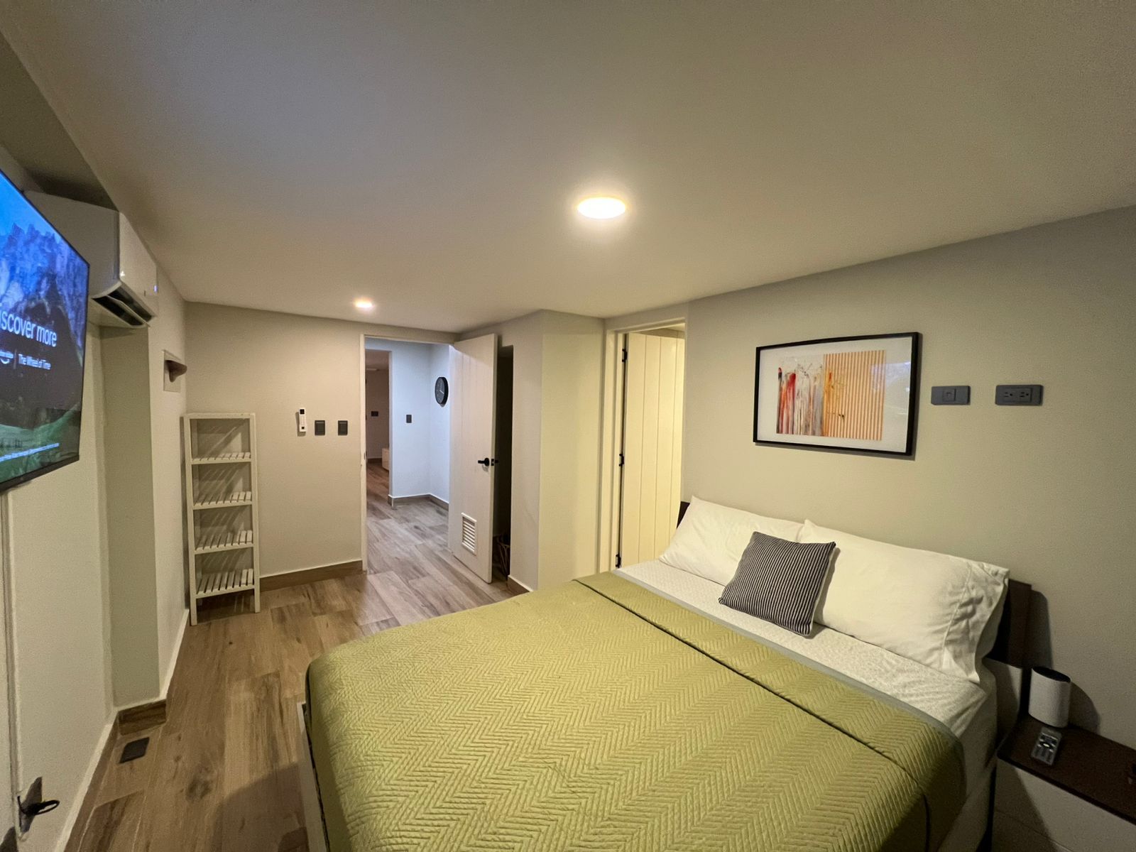 Habitació d'hotel amb un llit, televisió, quadre i un passadís que condueix a una altra habitació.