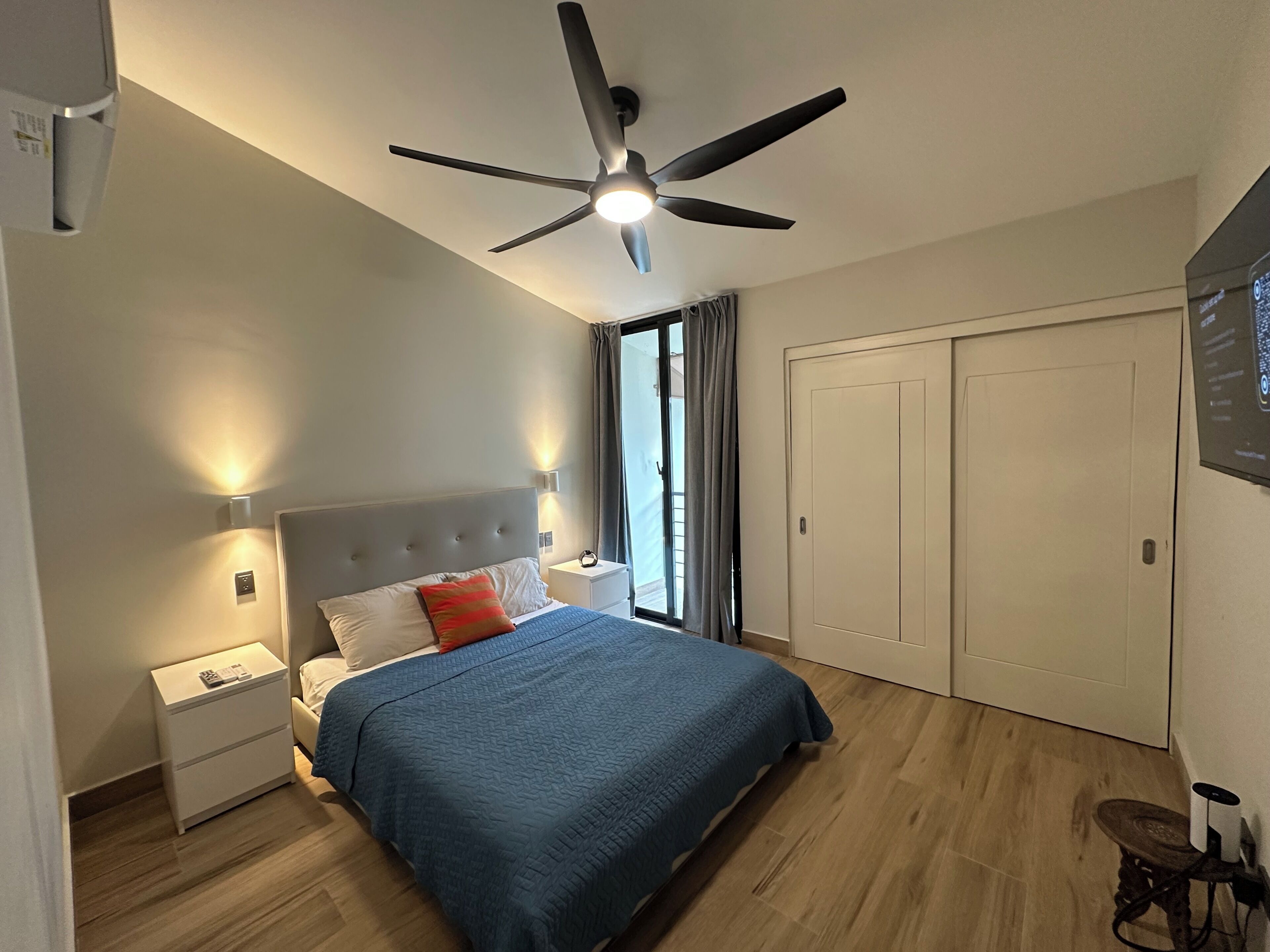 Een slaapkamer met een bed, nachtkastje, plafondventilator, raam met gordijnen en een deur.