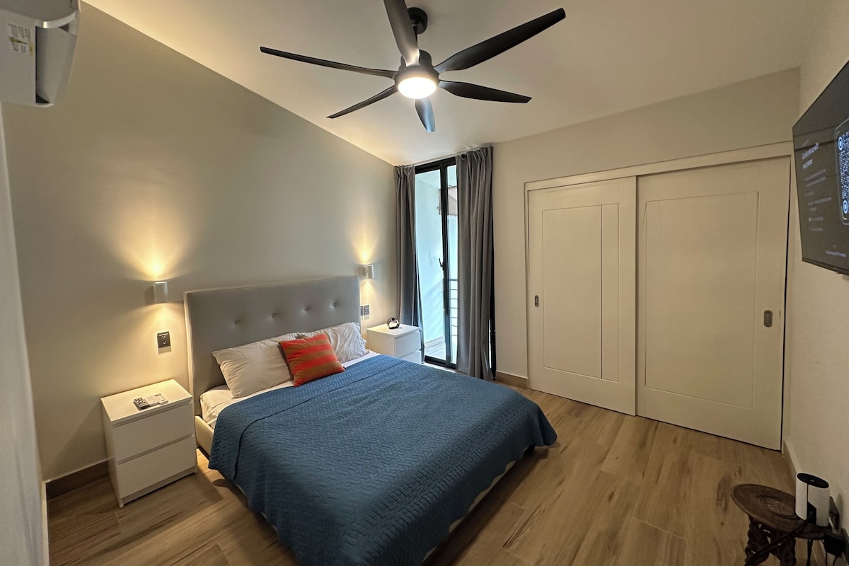 Un dormitorio con una cama, una mesita de noche, un ventilador de techo, una ventana con cortinas y una puerta.