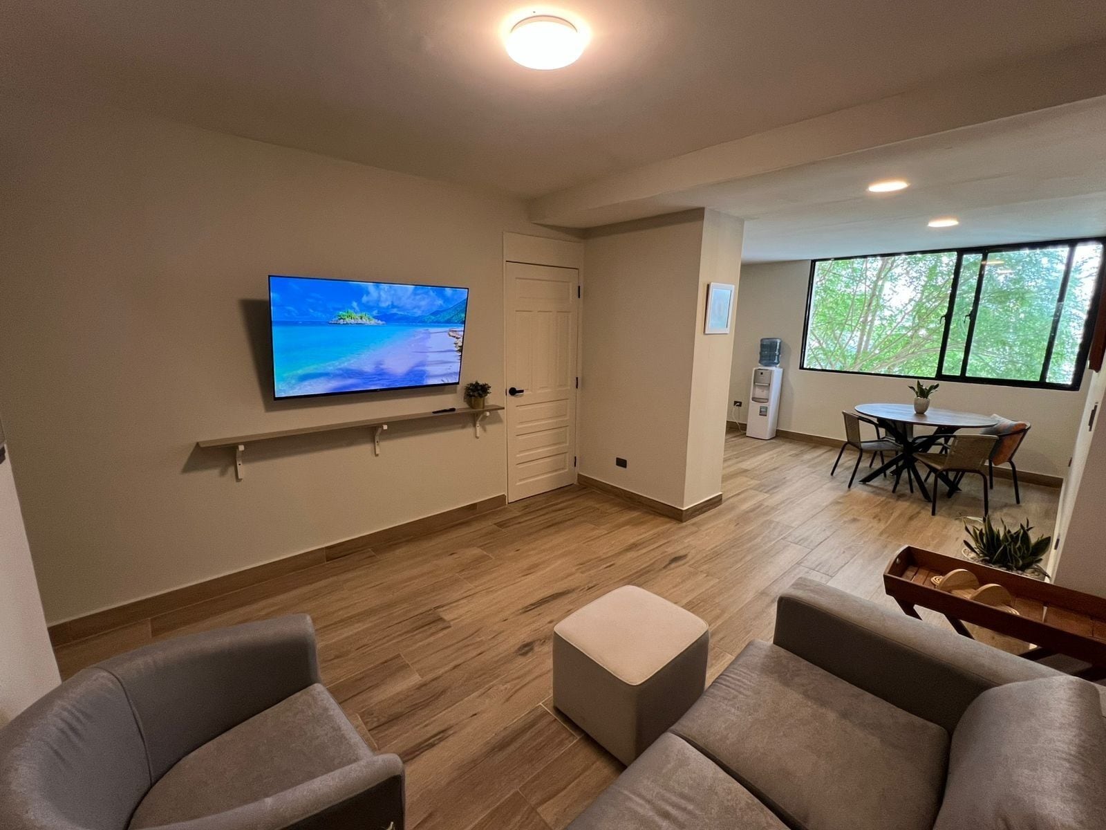 Ein modernes Wohnzimmer mit Fernseher, Essecke mit Tisch und Stühlen sowie einem großen Fenster mit Blick auf Grünflächen.