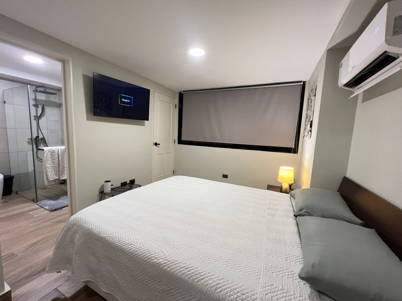 A hotel room with a bed, a television, an air conditioning unit, and a shower area.