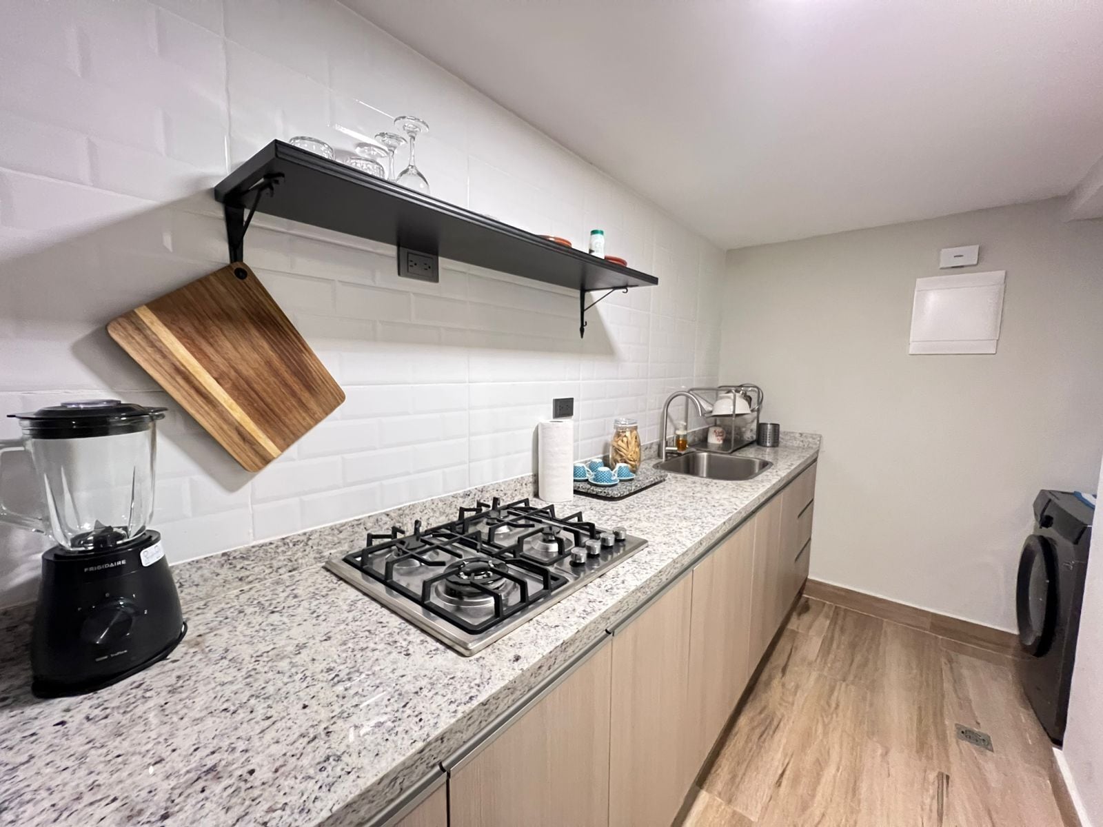 Une cuisine dotée d’un plan de travail en granit, d’une cuisinière à gaz, d’un mixeur, d’une planche à découper en bois, d’une étagère contenant de la verrerie et d’un évier.