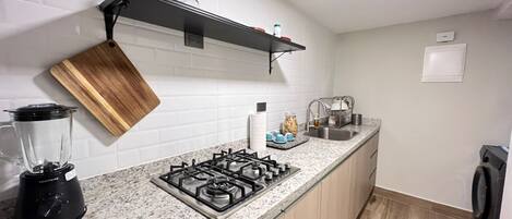 A kitchen with a granite countertop, a gas stove, a blender, a wooden cutting board, a shelf with glassware, and a sink.