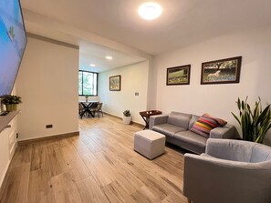 Ein modernes Wohnzimmer mit einem grauen Sofa, einem kleinen Hocker und einem Essbereich mit Tisch und Stühlen.