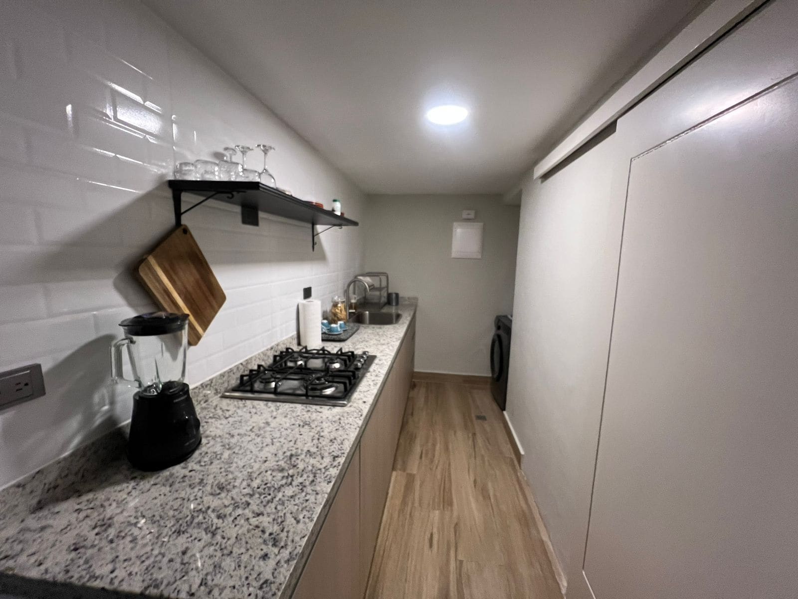 Une cuisine compacte équipée d’une cuisinière à gaz, d’un mixeur, d’une planche à découper en bois et d’une étagère où sont rangés des verres.