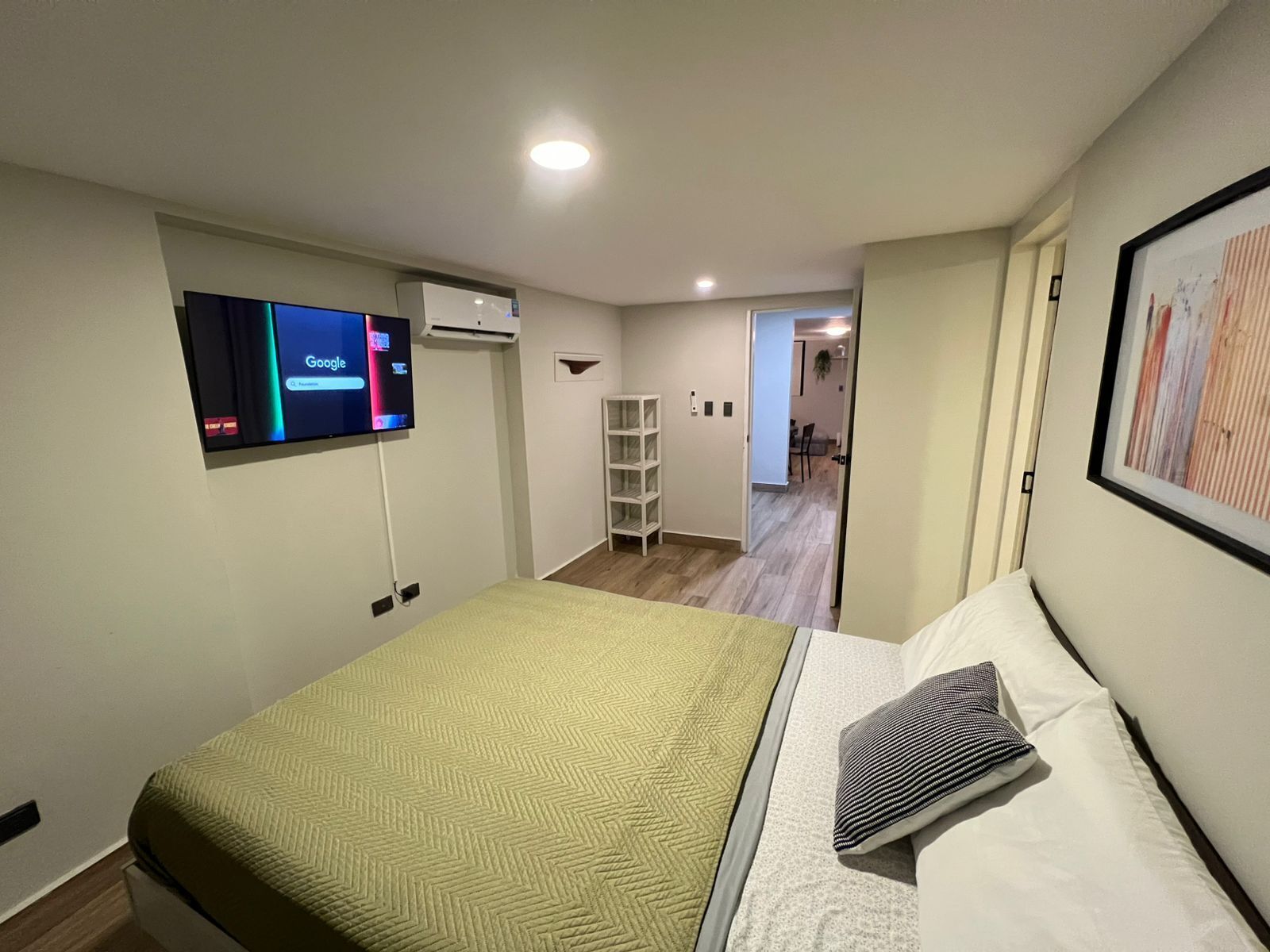 Quarto de hotel com cama, televisão fixada na parede, ar condicionado branco e corredor que dá para outro quarto.
