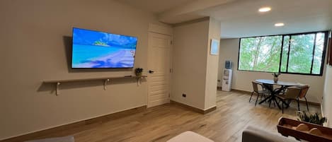 A modern living room with a flat-screen TV, a dining area with a table and chairs, and a large window with a view of trees.