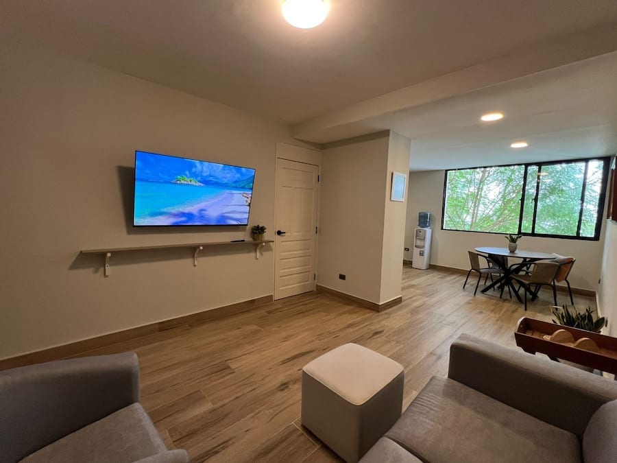 A modern living room with a flat-screen TV, a dining area with a table and chairs, and a large window with a view of trees.