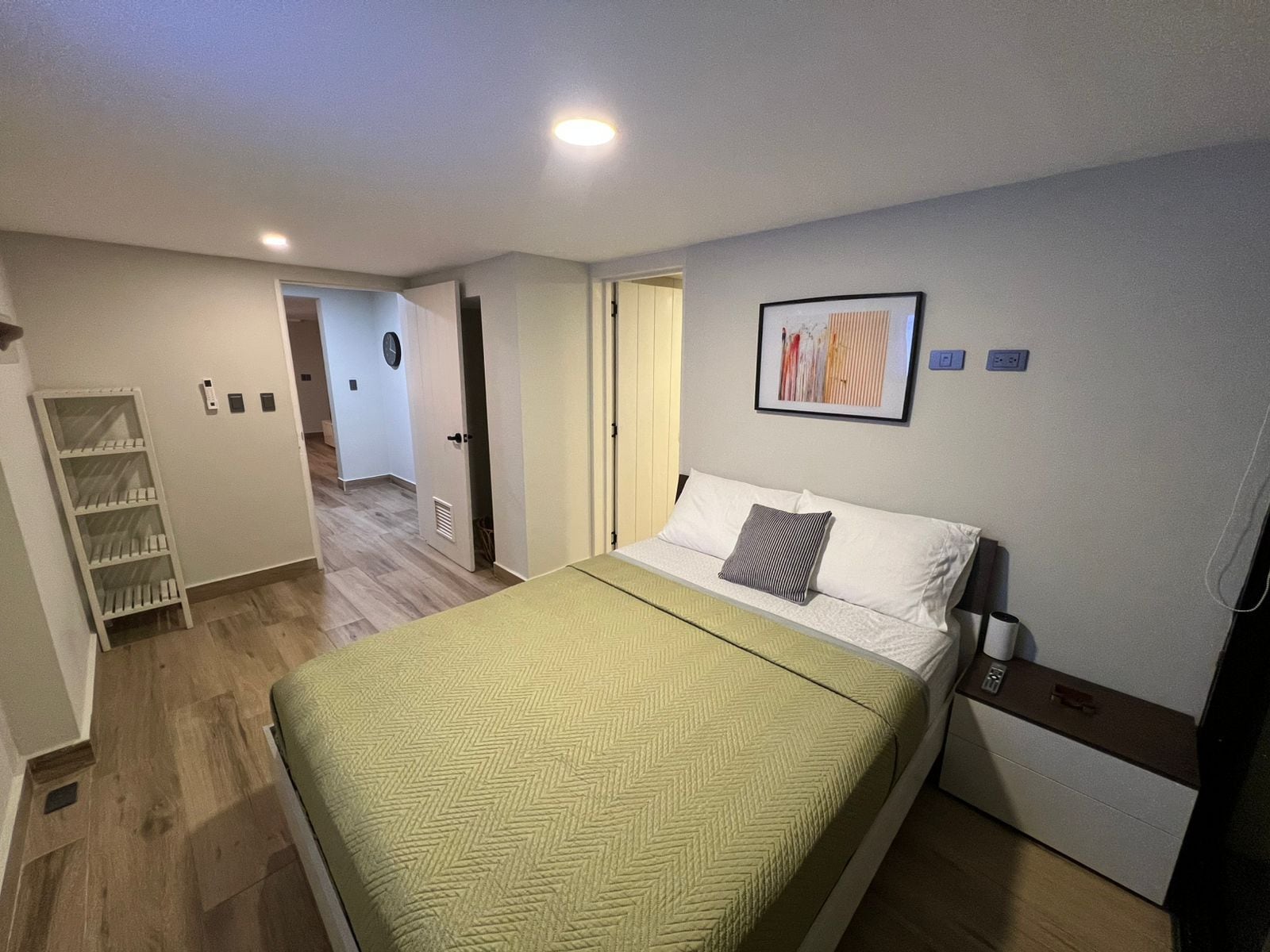 Quarto de hotel com cama, mesa de cabeceira, obra de arte na parede e porta que dá para outro cômodo.