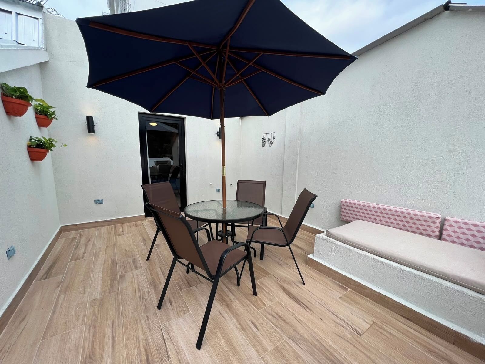 Een terras met een ronde tafel, stoelen, een grote parasol en een zachte zitplek.