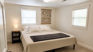 Quarto com cama, mesa de cabeceira, obra de arte na parede e duas janelas com persianas.