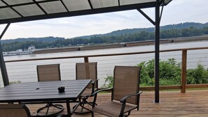 Uma área de estar coberta ao ar livre, com mesa e cadeiras, com vista para um rio, uma ponte e um barco ao longe.