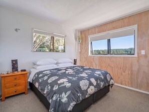A bedroom with a bed, a nightstand, a mirror, and a window with a view of trees. - Skippers View - Onemana Holiday Home (Whangamata)