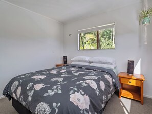 A bedroom with a bed, a nightstand, a window with a view of greenery, and a hanging plant. - Skippers View - Onemana Holiday Home (Whangamata)