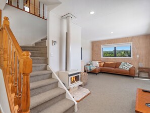 A staircase with wooden balusters, a fireplace with a visible fire, a brown leather sofa, and a window with a forest view. - Skippers View - Onemana Holiday Home (Whangamata)