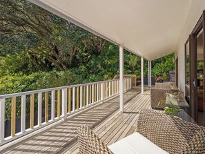 A covered outdoor deck with a wooden floor, white railings, wicker furniture, and a view of dense greenery. - Skippers View - Onemana Holiday Home (Whangamata)