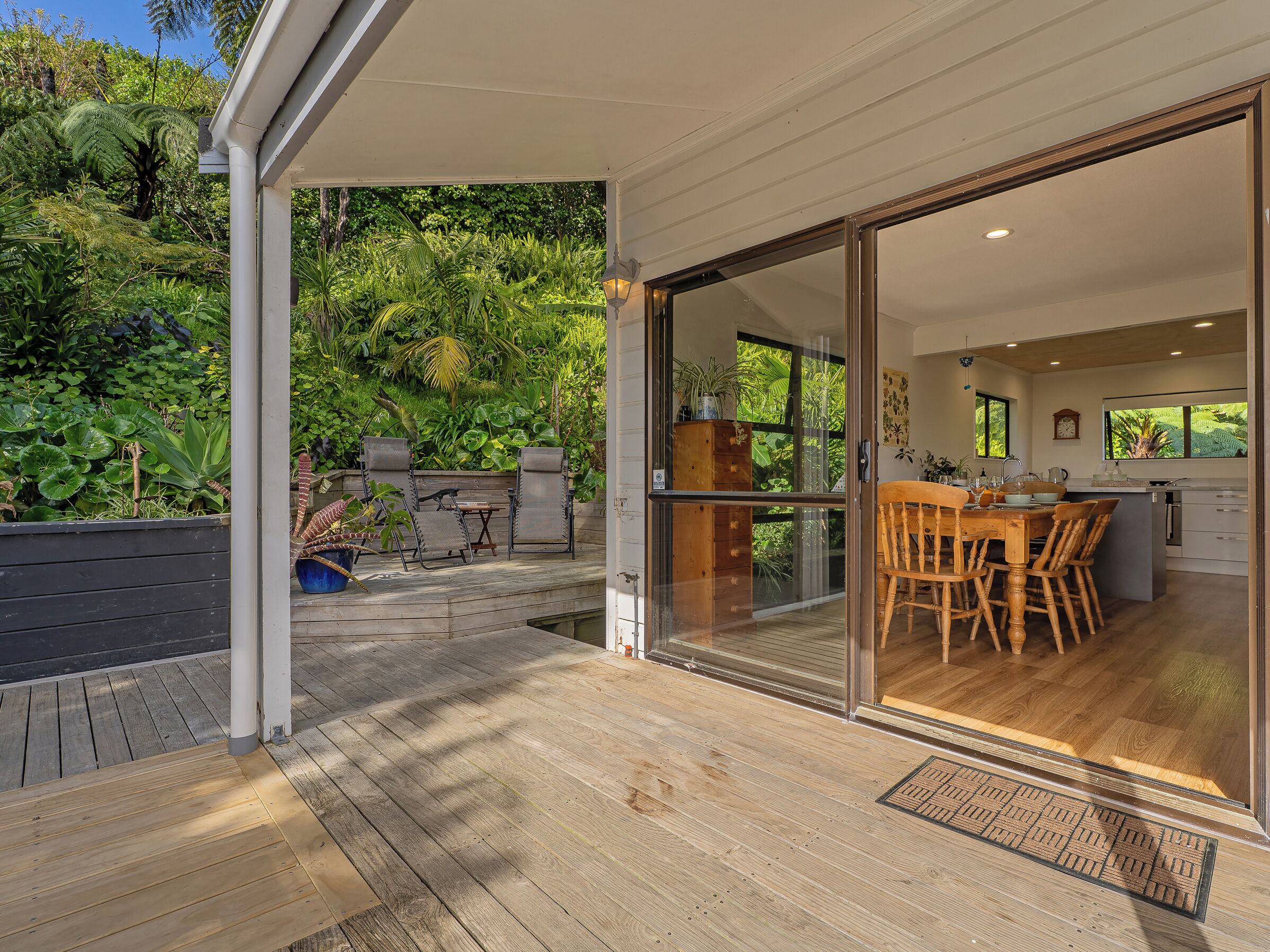 A covered outdoor area with a wooden deck, a dining table with chairs, and a view of a lush garden through glass doors leading to a kitchen area.