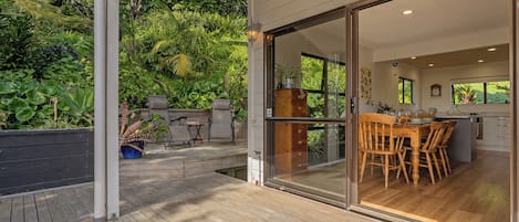 A covered outdoor area with a wooden deck, a dining table with chairs, and a view of a lush garden through glass doors leading to a kitchen area.