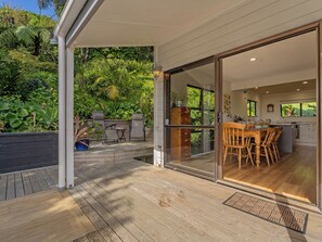 A covered outdoor area with a wooden deck, a dining table with chairs, and a view of a lush garden through glass doors leading to a kitchen area. - Skippers View - Onemana Holiday Home (Whangamata)