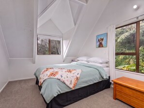A bedroom with a sloped ceiling, a bed with a floral bedspread, a wooden bench, and a window with a view of greenery. - Skippers View - Onemana Holiday Home (Whangamata)