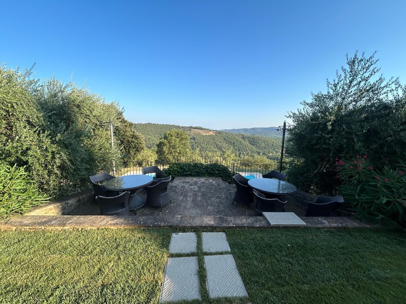Un espace extérieur aménagé avec deux tables rondes et des chaises, entouré d’une végétation luxuriante et offrant une vue sur des collines lointaines.