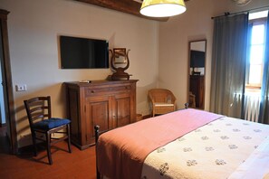 Quarto com cama, cômoda de madeira, cadeira, televisão na parede e janela com cortinas.
