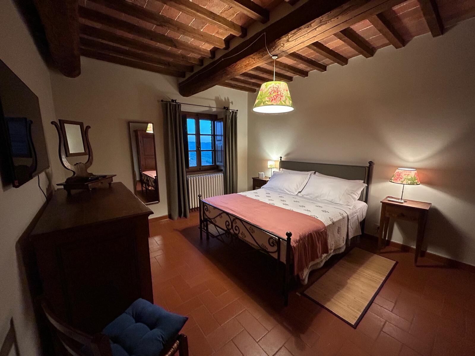 A bedroom with a bed, a chair, a desk, a mirror, a window with curtains, and a hanging lamp.