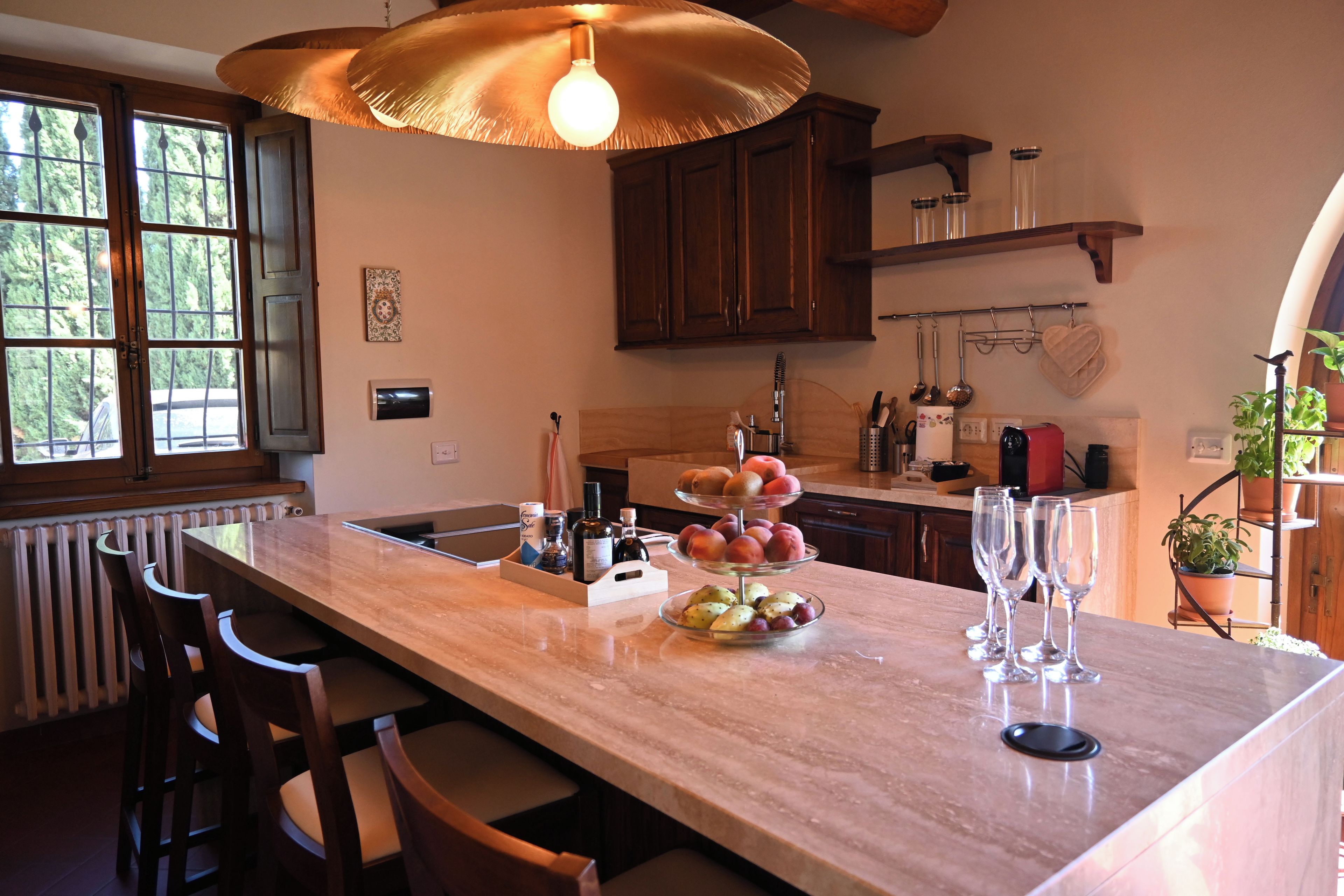 Una cocina con encimera de mármol, sillas de madera, lámpara colgante y ventana con vista a árboles.